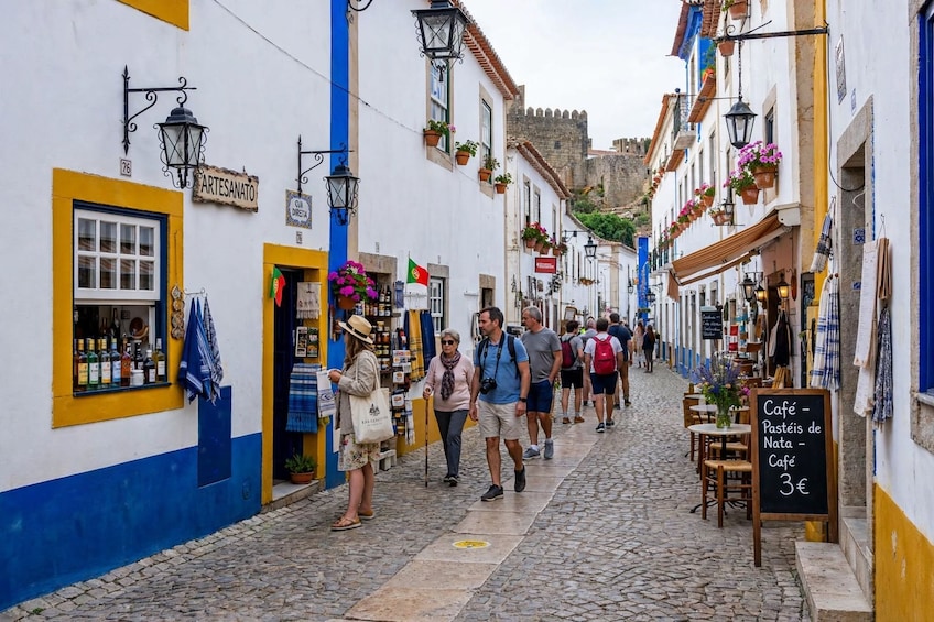 A cobblestone street lined with white buildings with blue and yellow accents, a sign for a café, and a castle in the background. 2