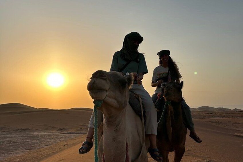 Two people riding camels on a desert road with the sun setting in the background. 4