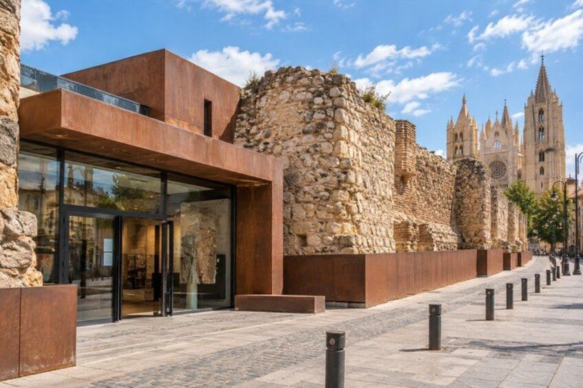 A modern building with large glass windows, a stone wall, and a cathedral in the background. 1