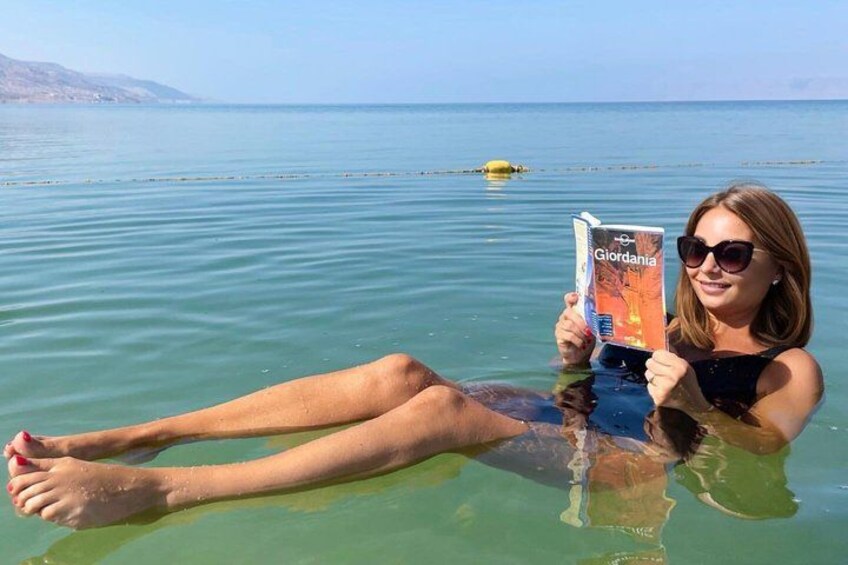 A person lying in calm water with a book titled 'Giordania,' a clear sky, and distant land. 1