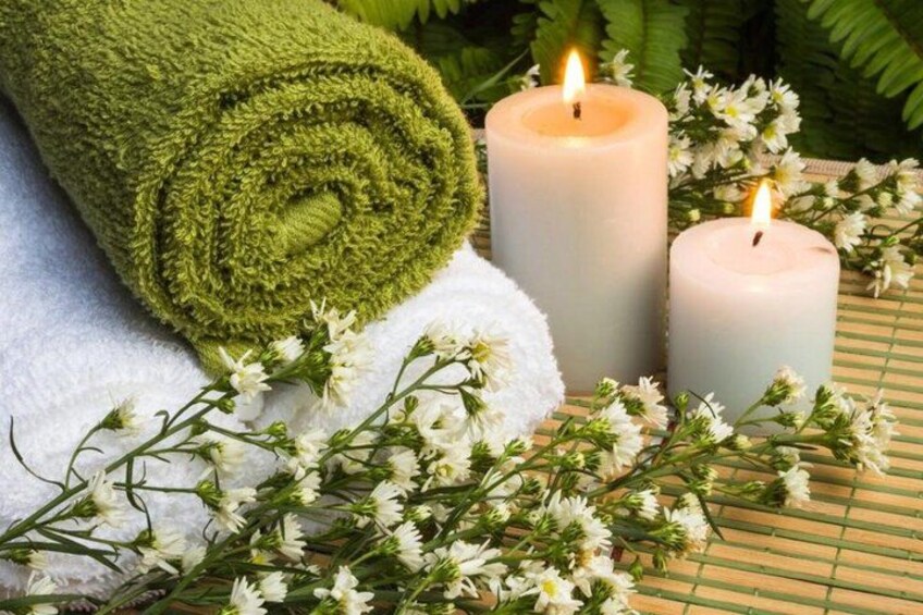 A rolled green towel, two lit candles, and white flowers on a bamboo mat. 4