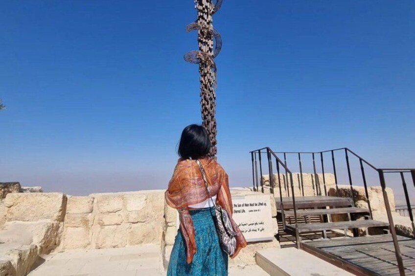 A person standing at a historical site with a tall, decorated column and a sign with text. 4