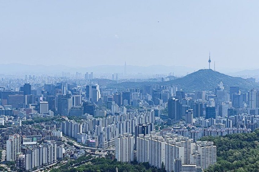 A densely populated cityscape with numerous high-rise buildings, a prominent hill with a tower, and a distant mountain range. 1