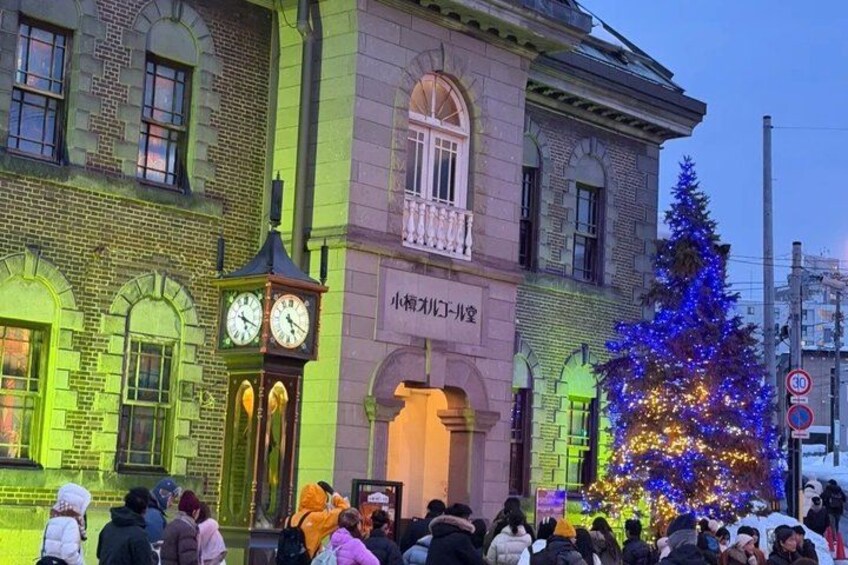 A building with a clock tower, a Christmas tree, and a crowd of people. 1