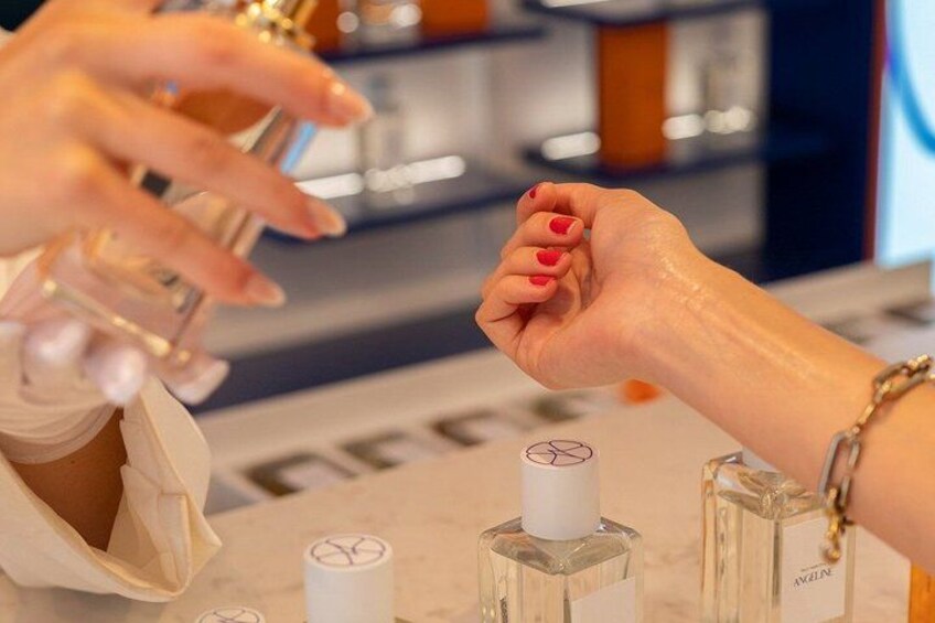 A person holding a perfume bottle with a white cap, and another hand with red nail polish reaching towards it. 5