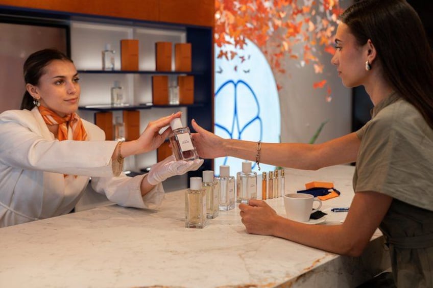 A person in a white coat handing a bottle to another person at a counter with various bottles and a large circular sign in the background. 1
