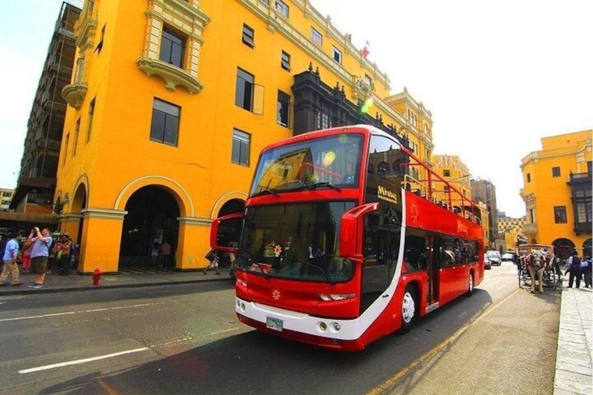 A red double-decker bus on a city street with yellow buildings in the background. 1
