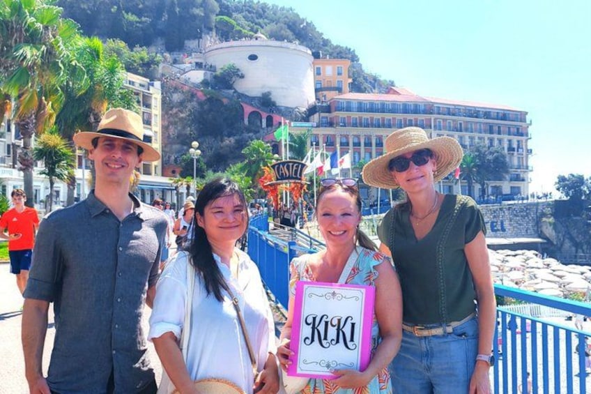 A group of people on a seaside promenade with a building and palm trees in the background. 1