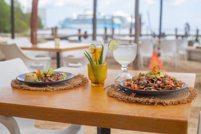 A wooden table with two plates of food and a glass of drink, set on a beach with chairs and umbrellas in the background. 2