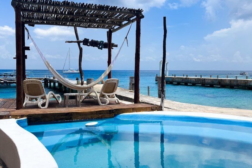 A pool with lounge chairs and a hammock under a wooden pergola by the sea. 1