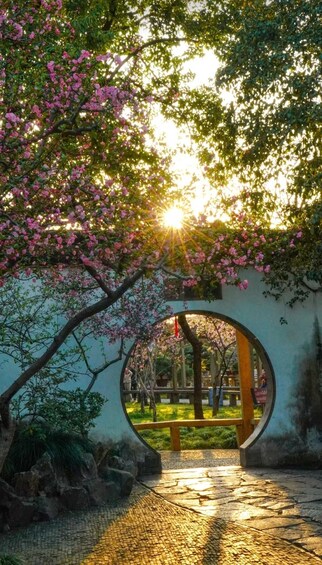 A stone pathway leading through a circular archway, surrounded by lush greenery and pink flowering trees. 3