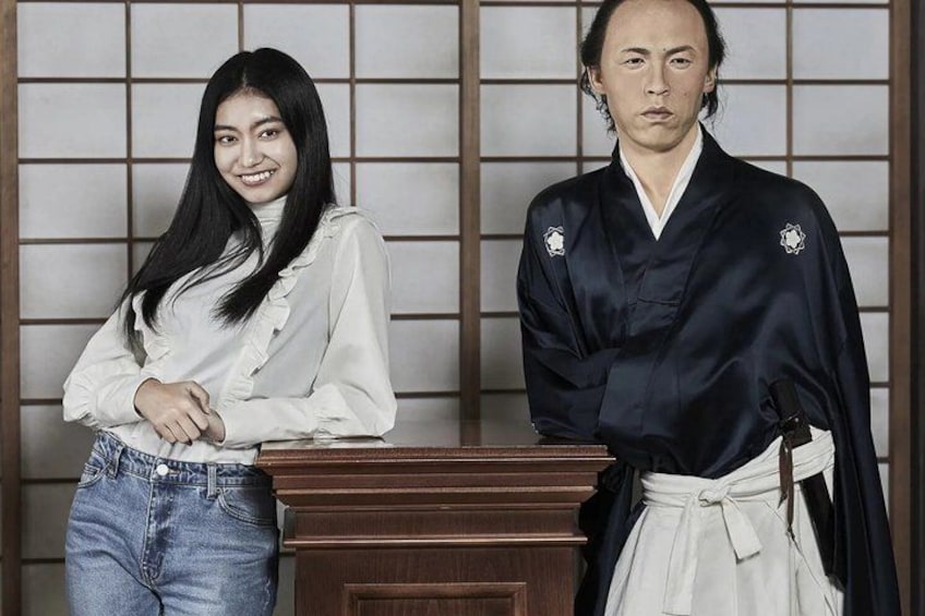 A person in a white blouse and jeans standing next to a wooden desk, and another person in traditional attire with a sash. 5