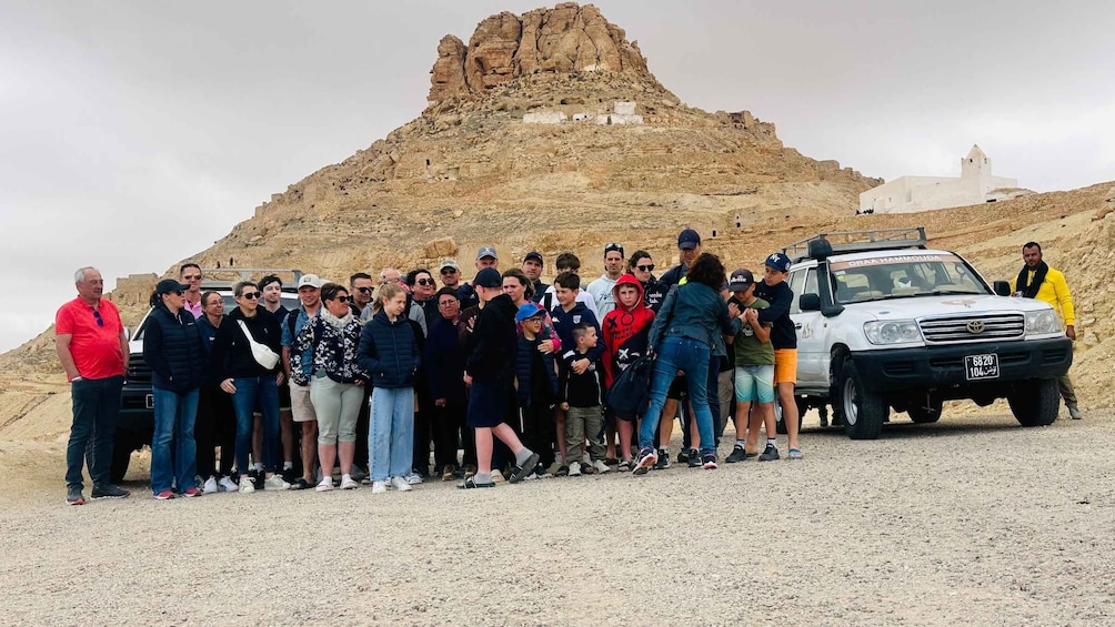 A group of people gathered in a rocky outdoor setting with a white vehicle parked nearby. 3