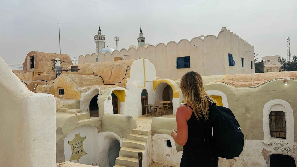 A person walking through a traditional village with white-washed buildings and a mosque with a minaret in the background. 1