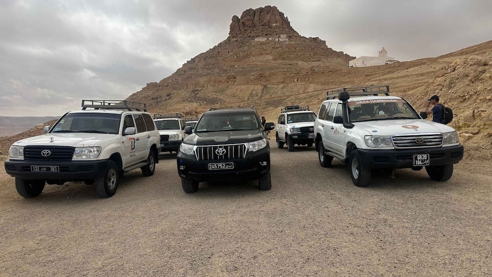 A convoy of white Toyota SUVs parked on a gravel road with a rocky hill and a white building in the background. 5