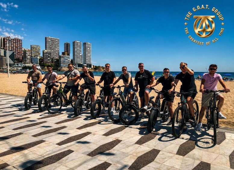 A group of people on bicycles on a tiled walkway by the beach with a city skyline in the background. 1