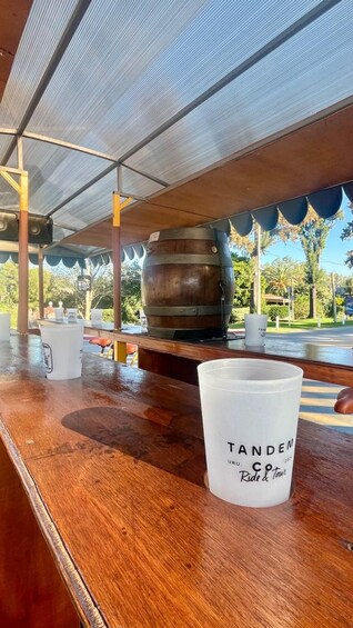 Een houten bar met een groot metalen vat, een wit kopje met de tekst 'TANDEM URU COFFEE Ride & Tour', en een overdekt buitengebied met bomen. 2