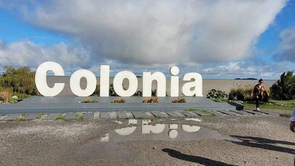 Een groot wit bord met de tekst 'Colonia' in het midden, omringd door een grindgebied met kleine bloemstukjes. 3