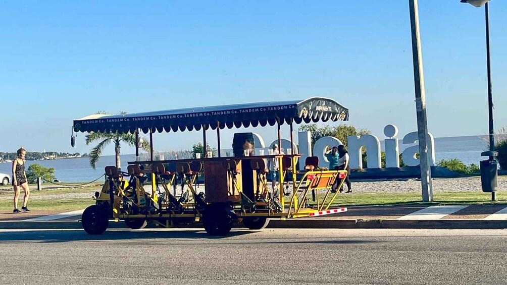 Een trolley met een blauw dak en 'TANDEM CO.' branding, geparkeerd op een straat met een helder uitzicht op de oceaan en een bord waarop 'Colonia' staat in de achtergrond. 1