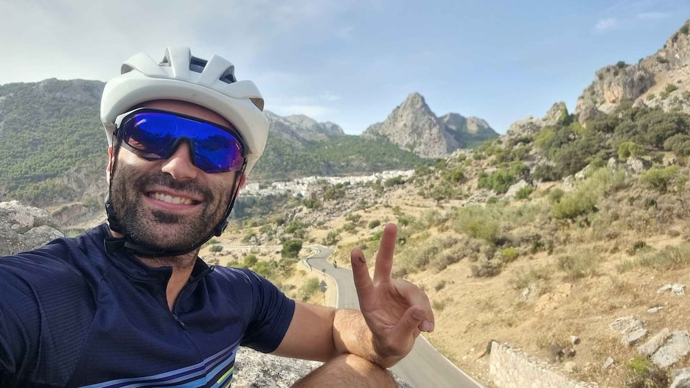 A person wearing a helmet and sunglasses, pointing towards a mountainous landscape with a road. 2