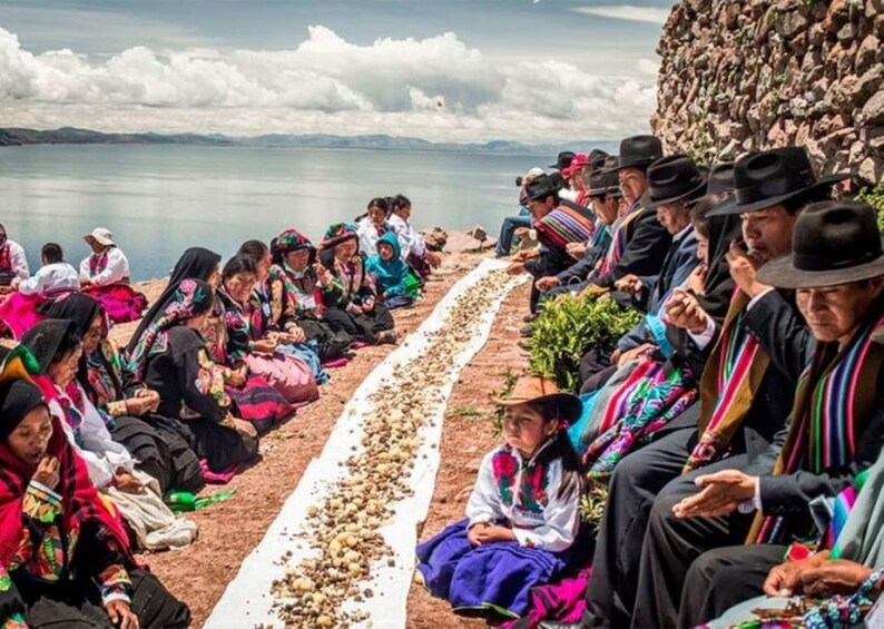 A group of people sitting on a dirt path by a body of water, dressed in colorful traditional clothing. 2