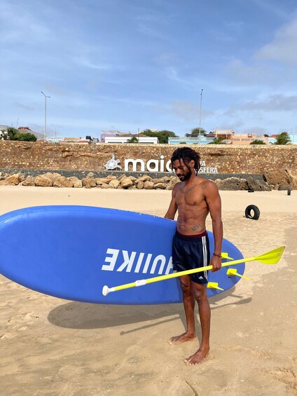 A man standing on a sandy beach holding a blue surfboard and a yellow paddle. 3
