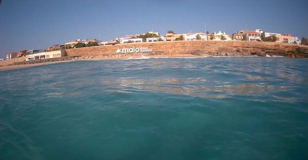 A coastal town with buildings on a cliff, clear blue sea, and a rocky shoreline. 2