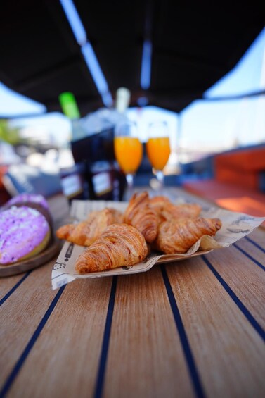 Una mesa de madera con cruasanes, un postre morado y dos vasos de jugo de naranja. 2