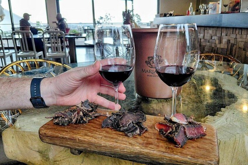 A person holding two glasses of red wine, a wooden board with food, and a branded container. 3