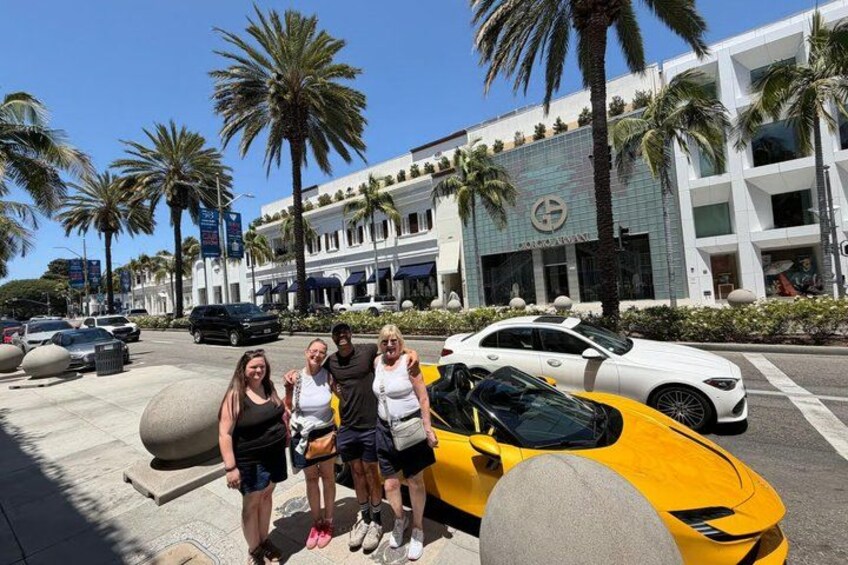 A group of people posing in front of a yellow sports car and a white luxury car on a sunny day. 2