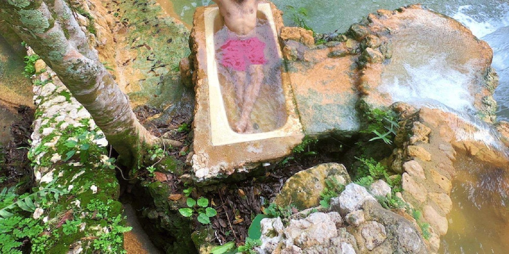 Eine Person in einer Steinschaukelbadewanne, umgeben von Steinen und Grünpflanzen. 4