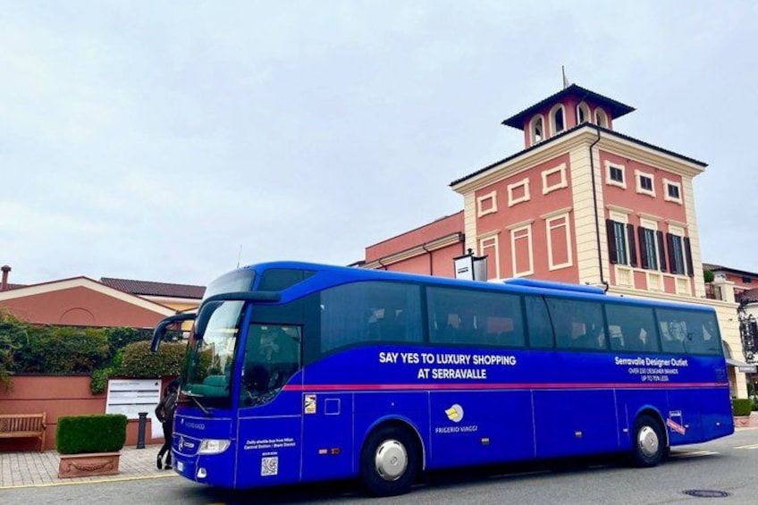 A blue bus with 'SAY YES TO LUXURY SHOPPING AT SERRAVALLE' on its side, parked in front of a building with a red and beige facade. 1