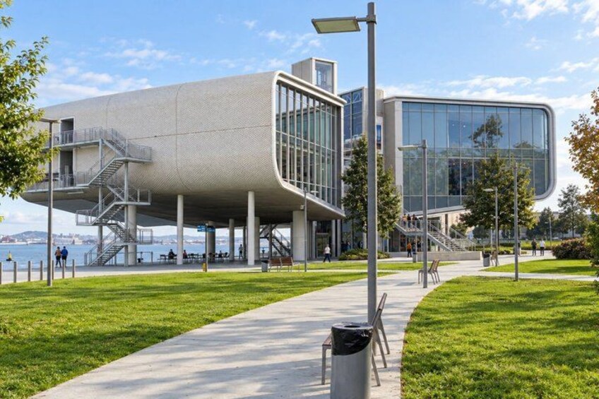A modern building with a unique curved design, a waterfront view, and a pathway leading to it. 4