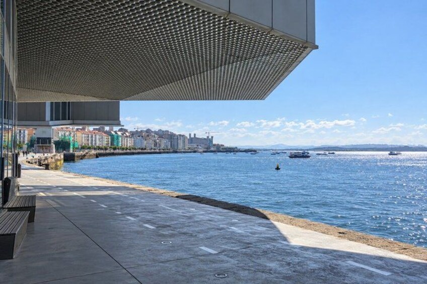 A waterfront promenade with a modern building, a clear sky, and boats on the water. 2