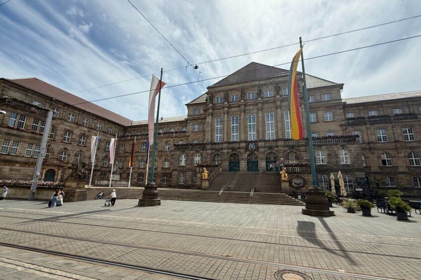A large historic building with multiple flags, a central staircase, and a paved plaza. 5