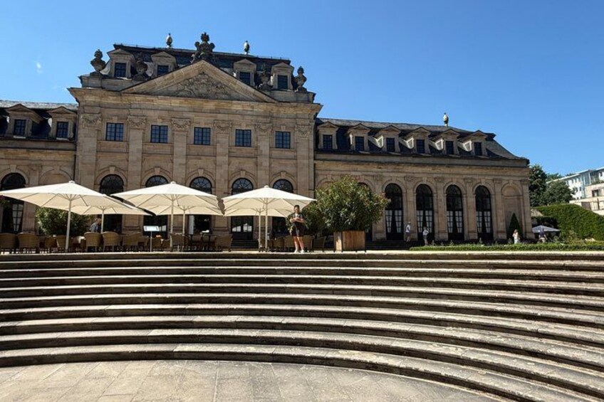 A grand building with classical architecture, a set of stairs leading up to it, and outdoor seating with umbrellas. 2
