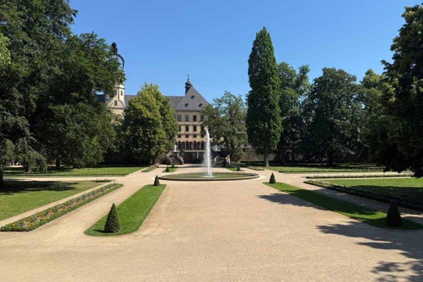A formal garden with a fountain, manicured lawns, and a large building in the background. 1