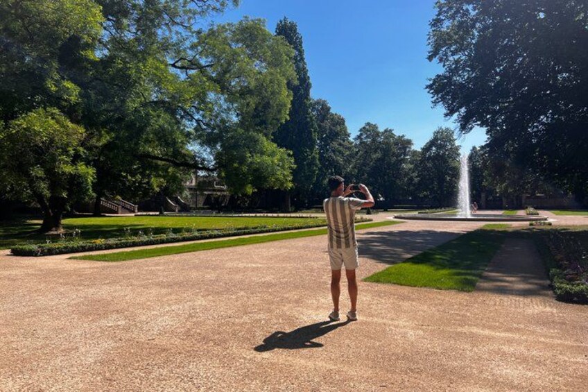 A person taking a photo of a fountain in a park with trees and a well-maintained lawn. 3