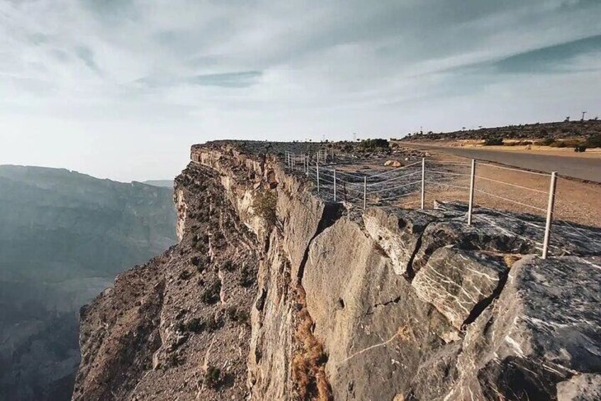 A rocky cliff edge with a guardrail, a road in the background, and a hilly landscape. 5