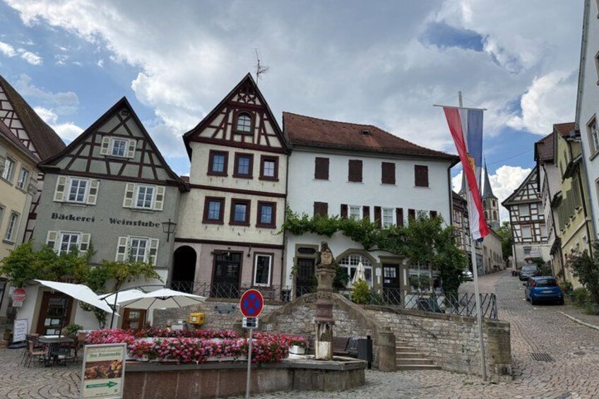 A cobblestone street with traditional half-timbered houses, a sign for a bakery, and a flagpole with flags. 2