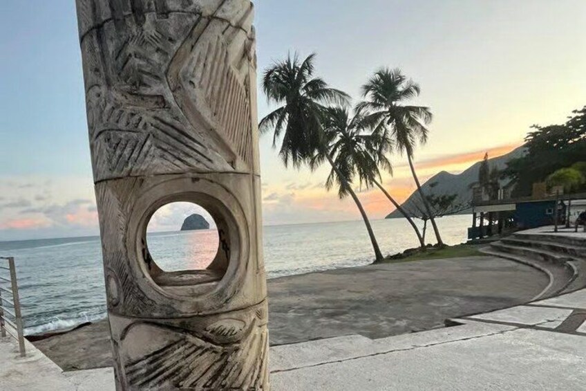 A carved tree trunk with a circular window, palm trees, a beach, and a mountain in the background. 1