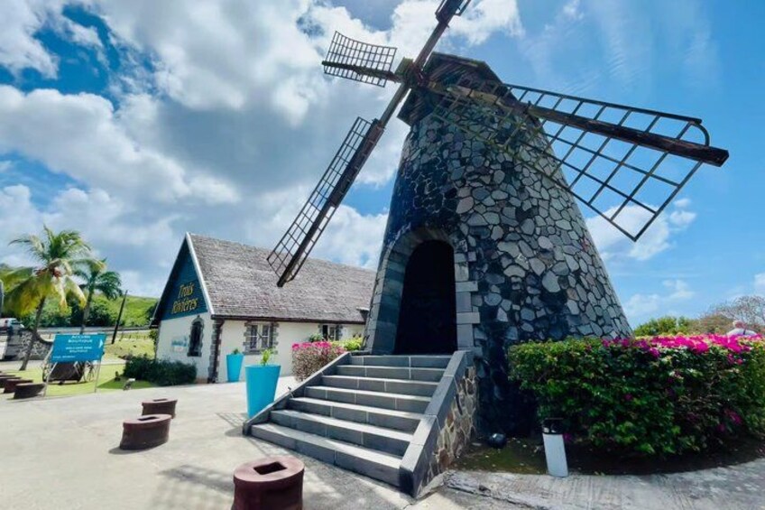 A stone windmill with a staircase leading up to its entrance, a small building with a sign, and lush greenery. 4