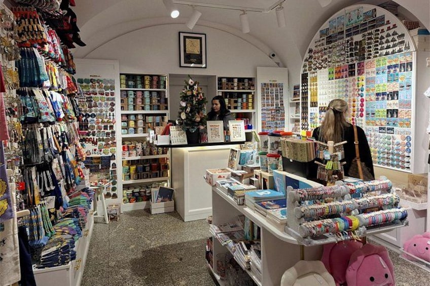 A gift shop with shelves of various items, a counter with a person behind it, and a wall with hanging items. 2