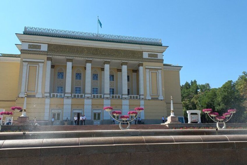 A large building with columns and a flag, surrounded by a fountain with pink flowers. 1