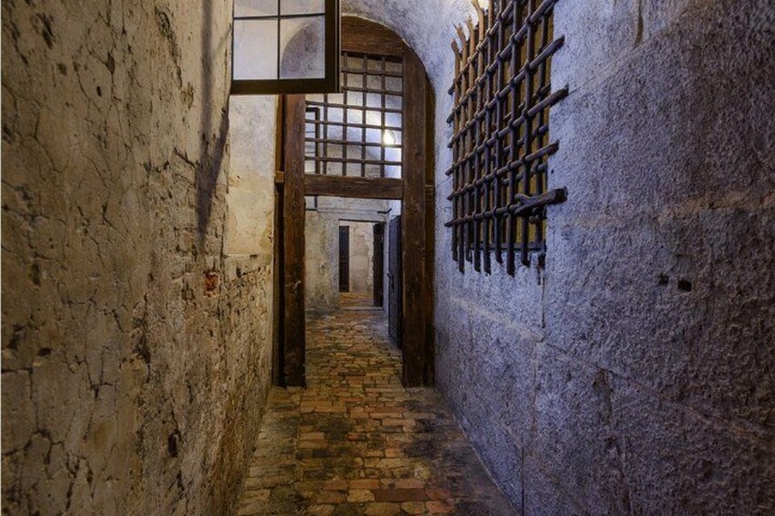 An arched stone corridor with a brick floor, a wooden door, and a barred window. 4