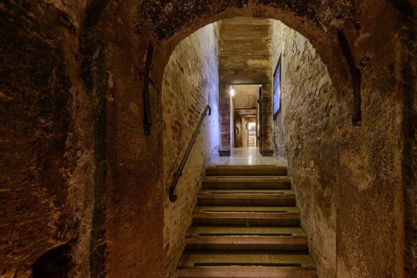 A stone staircase in a tunnel with a metal handrail and a light at the end. 3