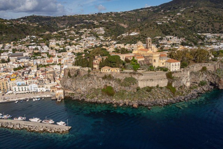 A coastal town with a historic fort, clear blue waters, and a hilly backdrop. 1