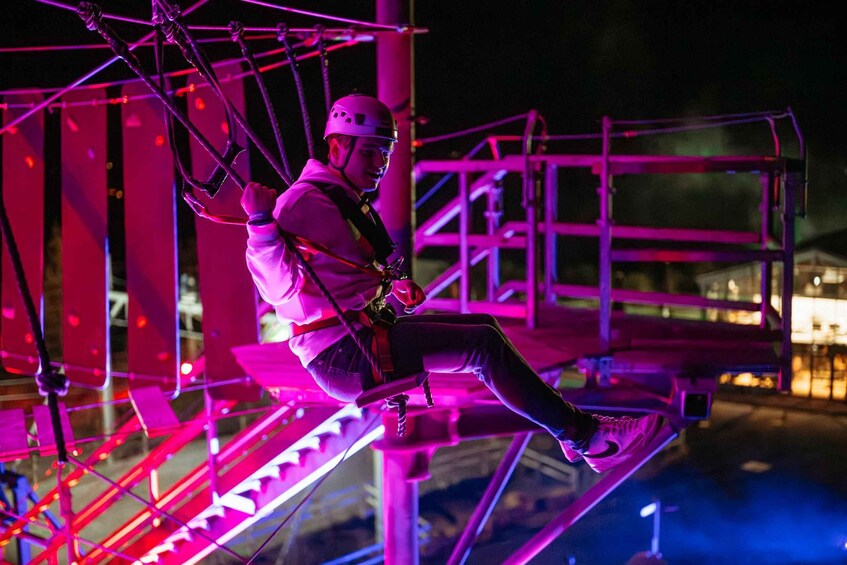 A person on a zip line with a safety harness, suspended between metal structures with a purple-lit background. 4