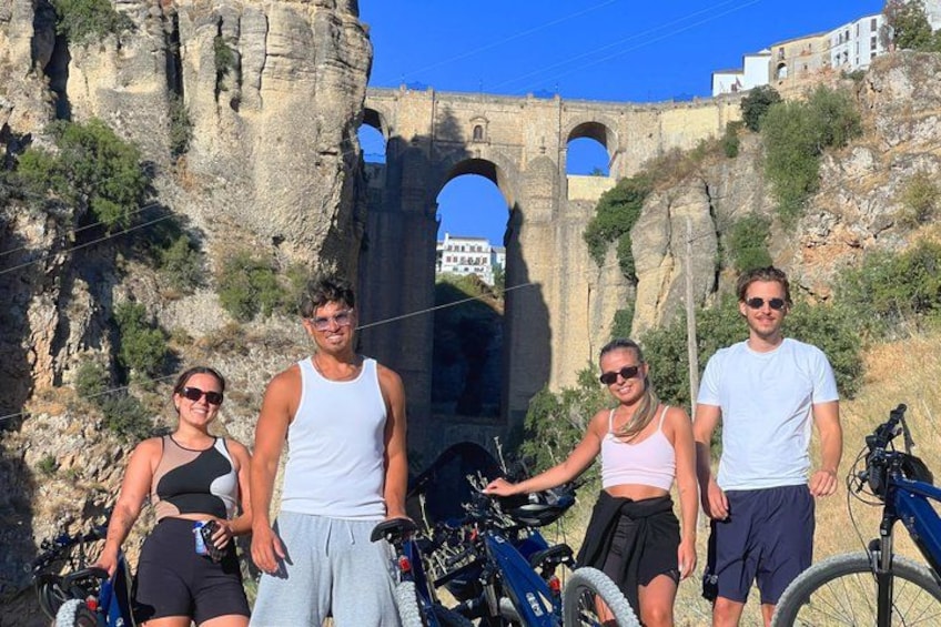 A group of people standing in front of a historical stone bridge with arches, surrounded by rocky terrain and greenery. 1