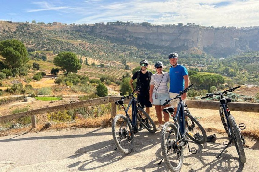 Three people standing on a paved path with bicycles, overlooking a hilly landscape with trees and cliffs. 5